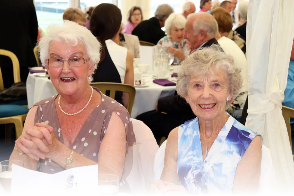 two ladies sat in a busy function room, smartly dressed and smiling for the camera