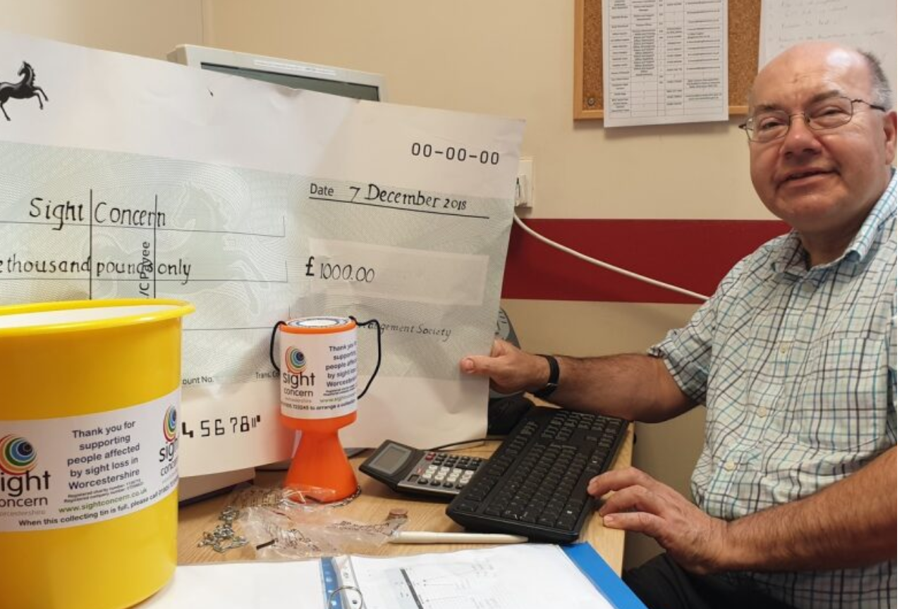 a man sat at a desktop holding an over-sized cheque of £1000 paid to Sight Concern. there are collections buckets on the desk.